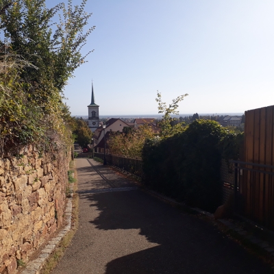 Depuis le haut d'une rue, vue sur le clocher d'une église aux murs blancs et au toit très pointu, très élancé. Un mur de briques à gauche, des maisons à droite